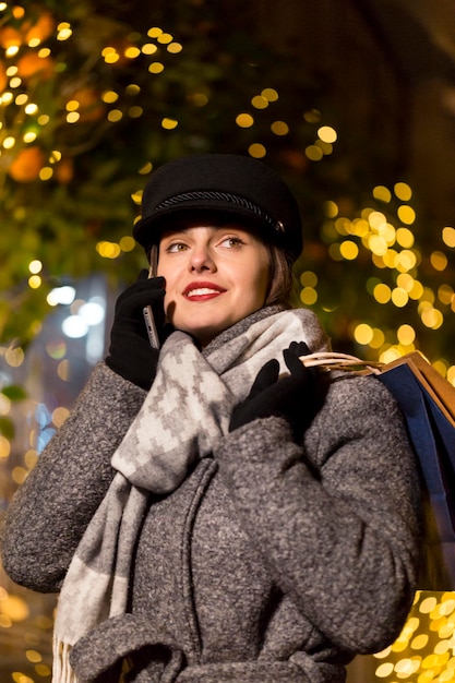 Elegante chica morena sonriente sosteniendo bolsas de compras y un teléfono inteligente antes de la pared de guirnaldas en la noche de Navidad de la ciudad