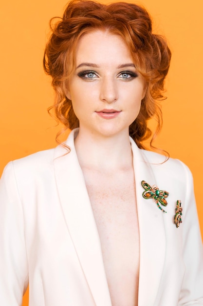 Elegante chica pelirroja posando sobre un fondo naranja en una chaqueta blanca con un broche en forma de libélula