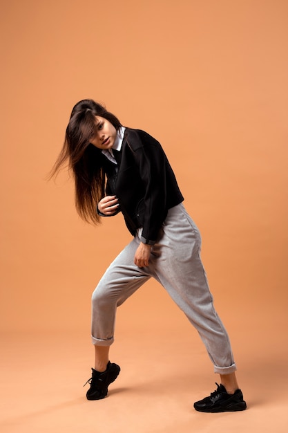 Elegante chica de pelo oscuro vestida con camisa blanca, pantalón gris, chaqueta negra y zapatillas negras está posando sobre el fondo beige en el estudio.