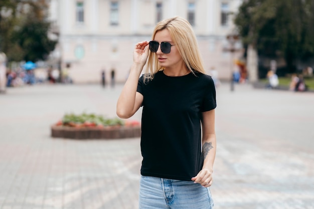 Elegante chica rubia con camiseta negra y gafas posando contra la calle