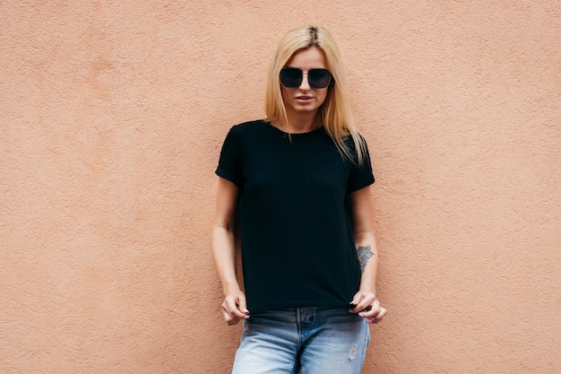 Elegante chica rubia con camiseta negra y gafas posando contra la pared