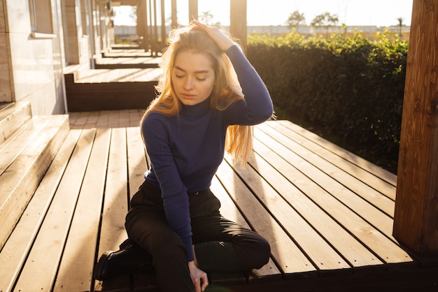 Elegante chica rubia de pelo largo en suéter azul se sienta en el parque bajo el sol
