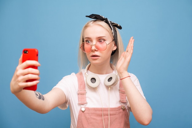 Foto elegante chica sorprendida en una brillante ropa casual y gafas de color rosa se alza sobre un fondo azul con un teléfono inteligente en sus manos y se ve sorprendida por la pantalla del teléfono