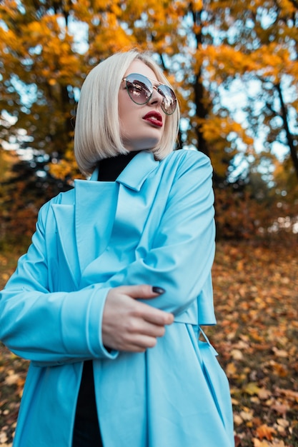 Elegante y colorido retrato de una hermosa mujer de moda con gafas de sol glamour en un abrigo azul en un bosque de otoño con follaje de otoño amarillo