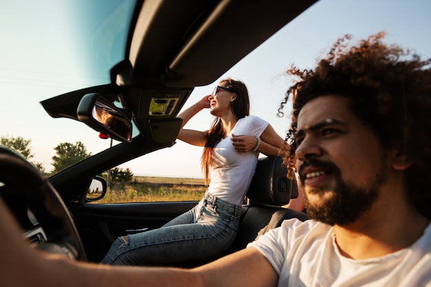 La elegante compañía de chicos y chicas jóvenes está sentada y sonriendo en un descapotable negro en la carretera en un día soleado. .