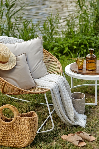 Elegante composición de jardín al aire libre en el lago con sillón de ratán de diseño, mesa de café, cuadros, almohadas, bebidas y elegantes accesorios.