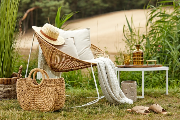 Elegante composición de jardín al aire libre en el lago con sillón de ratán de diseño, mesa de café, cuadros, almohadas, bebidas y elegantes accesorios.