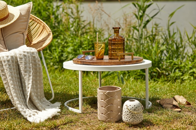 Elegante composición de jardín al aire libre en el lago con sillón de ratán de diseño, mesa de café, cuadros, almohadas, bebidas y elegantes accesorios.