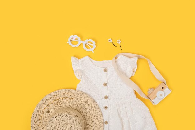 Elegante conjunto de ropa de verano para niños Vestido blanco, sombrero de paja, gafas de sol y accesorios sobre fondo amarillo Concepto de lookbook de chica de moda Vista superior Copiar espacio Plano