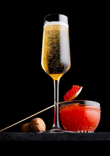 Elegante copa de champán amarillo con caviar rojo en cuchara de oro y recipiente de vidrio de caviar en tablero de mármol en negro.