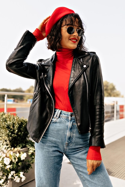 Elegante dama encantadora con gorra, chaqueta oscura y gafas de sol sonríe mientras está de pie en la ciudad en un día cálido y soleado