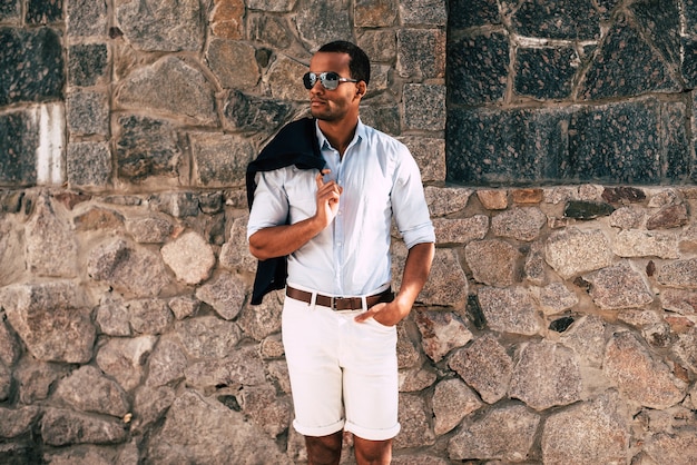 Elegante e bonito. Jovem africano bonito em roupas elegantes e casuais, carregando uma jaqueta no ombro e olhando para o lado de pé contra a parede de pedra ao ar livre