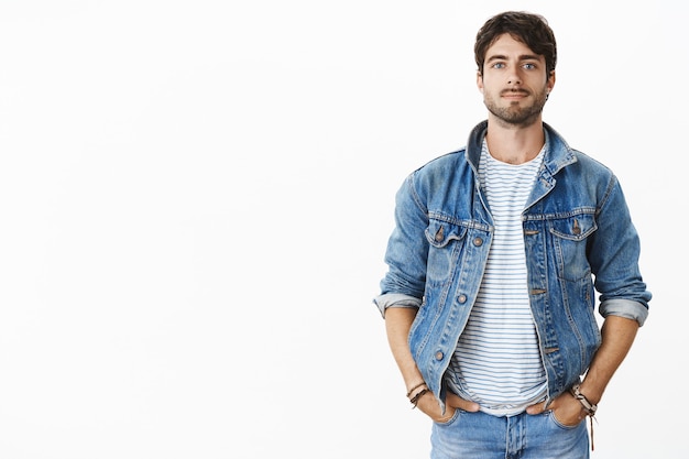 Elegante e cofnident jovem bonito com barba e olhos azuis de mãos dadas nos bolsos em pé contra uma parede cinza em uma jaqueta jeans da moda sorrindo alegremente esperando perto do carro pela namorada