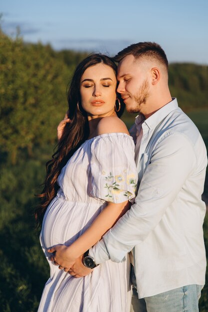 Elegante e elegante mulher grávida e seu marido vestiram um tom branco e azul pastel no campo por do sol