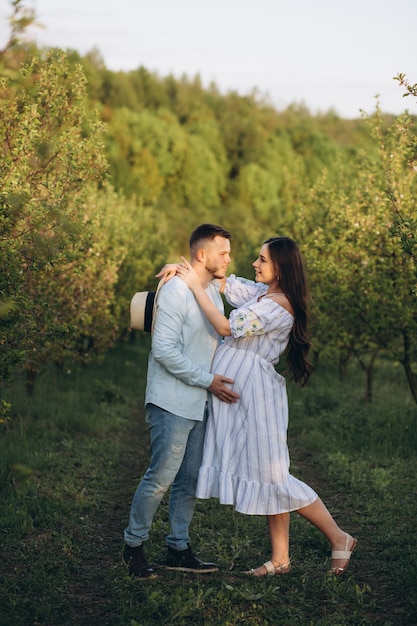 Elegante e elegante mulher grávida e seu marido vestiram um tom branco e azul pastel no jardim por do sol