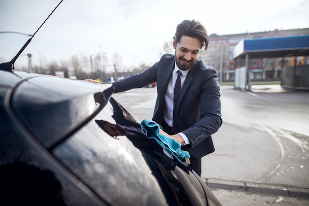 Elegante elegante alegre joven barbudo en un traje de limpieza de la ventana trasera del coche con un paño de microfibra azul.