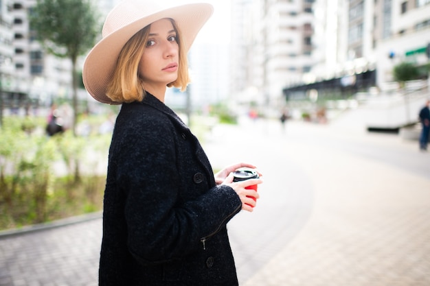 Elegante y elegante chica rubia de pelo corto con sombrero posando con café sobre fondo de calle