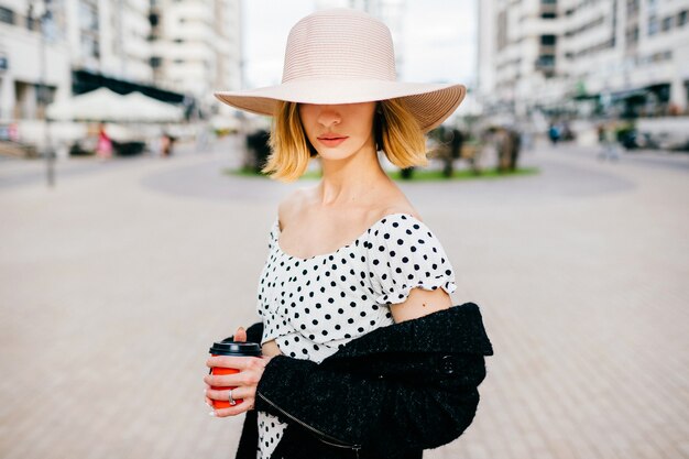 Elegante y elegante chica rubia de pelo corto con sombrero y vestido posando sobre fondo de calle