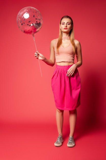 Elegante y elegante modelo de mujer joven con maquillaje brillante con top rosa y falda sosteniendo un globo transparente con confeti mientras está de pie contra el fondo rojo.