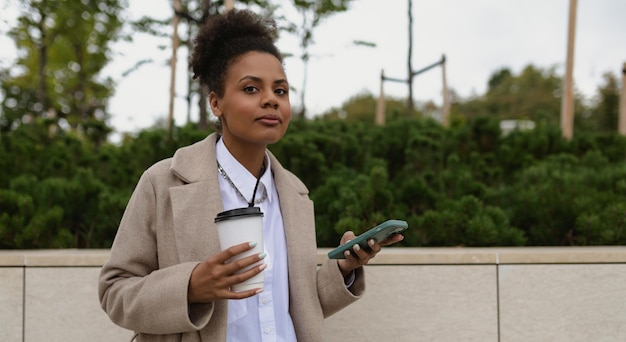 Elegante empresária afro-americana com uma xícara de café e um telefone nas mãos do lado de fora