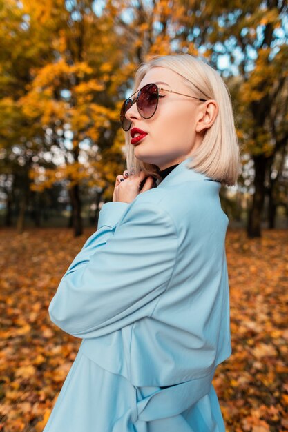 Elegante empresaria hermosa con gafas de sol de moda en un abrigo azul de moda camina sobre el fondo de follaje otoñal amarillo en el parque