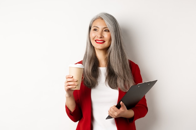 Elegante empresaria madura en chaqueta roja con descanso, sosteniendo el portapapeles y tomando café, mirando a la izquierda con una sonrisa complacida, fondo blanco.