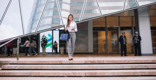 Elegante empresaria navegando por teléfono inteligente mientras está de pie en las escaleras contra el centro de negocios