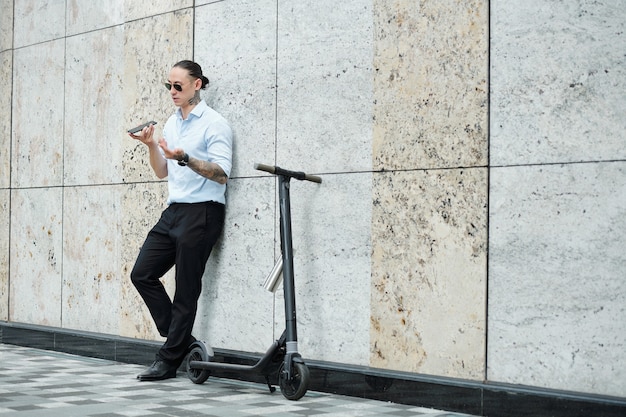 Elegante empresario apoyado en la pared del edificio cuando está de pie junto al scooter y graba un mensaje de voz