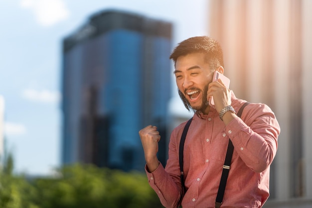 Elegante empresário asiático gesticulando sucesso enquanto fala com o celular