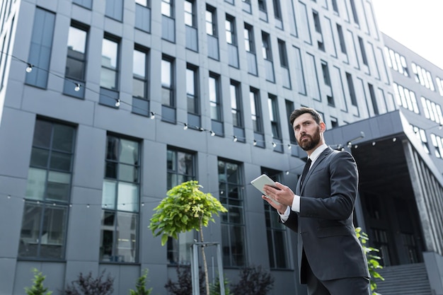 Elegante empresario barbudo en traje formal de pie trabajando con tableta en manos sobre fondo moderno edificio de oficinas exterior. Hombre que usa el teléfono inteligente o usa el teléfono móvil al aire libre en las calles de la ciudad