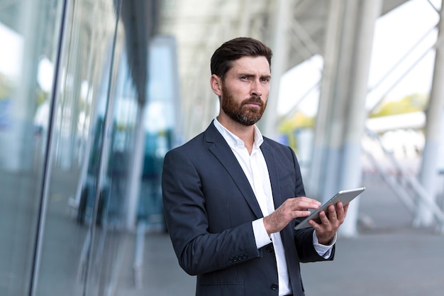 Elegante empresario barbudo en traje formal de pie trabajando con tableta en manos sobre fondo moderno edificio de oficinas exterior. Hombre que usa el teléfono inteligente o usa el teléfono móvil al aire libre en las calles de la ciudad