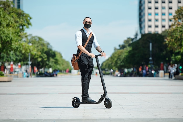 Elegante empresario guapo en máscara médica disfrutando de montar scooter eléctrico en la ciudad