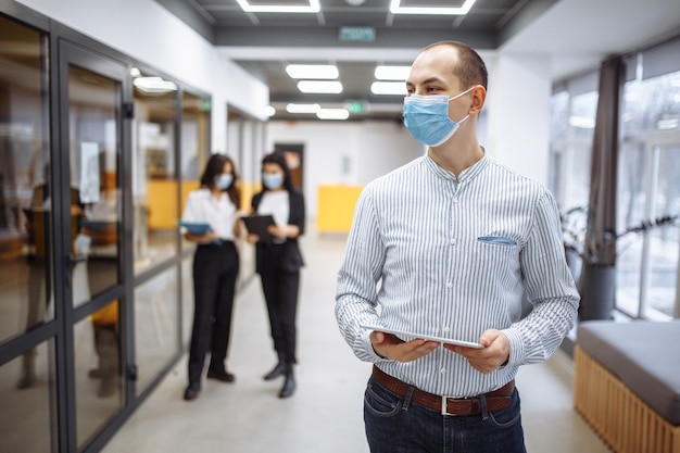 Elegante empresario con una máscara médica se encuentra en el pasillo de la oficina con una tableta en sus manos.