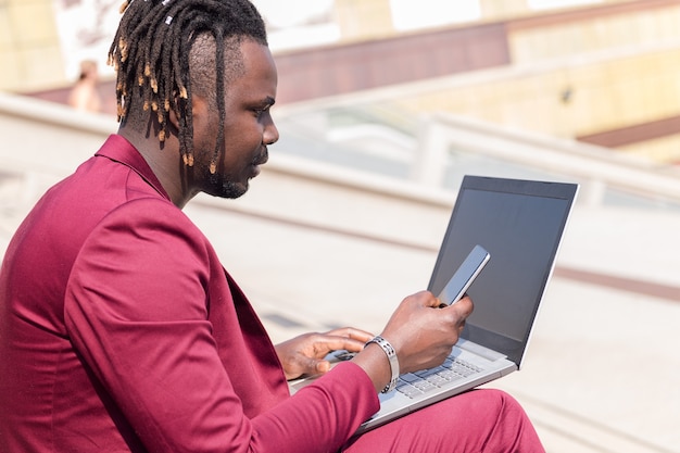 Elegante empresário negro trabalha ao ar livre com laptop e telefone, tecnologia e conceito de trabalho remoto