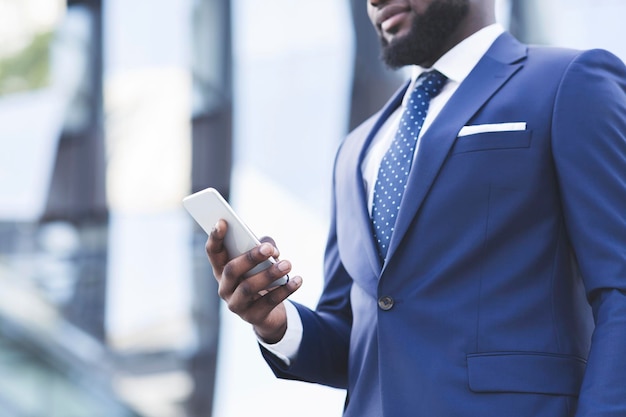 Elegante empresário negro usando smartphone caminhando ao ar livre cortado