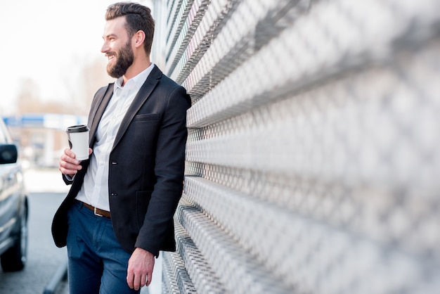 Elegante empresario de pie con café para ir cerca del coche en la moderna gasolinera