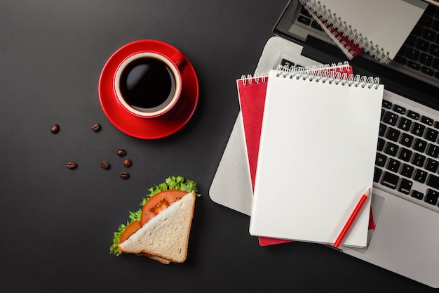 Elegante escritorio de oficina negro con laptop, taza de café y un sándwich para el almuerzo. Vista superior con espacio de copia.