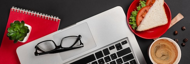 Elegante escritorio de oficina negro con laptop, taza de café y un sándwich para el almuerzo. Vista superior con espacio de copia.