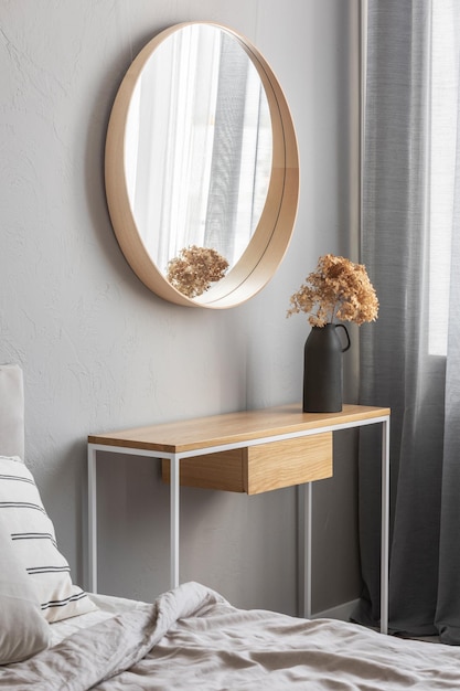 Elegante espejo redondo en marco de madera por encima de la mesa de la consola de fantasía con flores en el jarrón en el interior del dormitorio de moda con jarrón beige