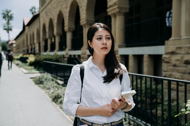 elegante estudiante de intercambio que sostiene un libro escolar que va a un curso en la universidad. colegiala trabajadora caminando al aire libre en stanford. una joven seria estudia en el extranjero solo en Estados Unidos.