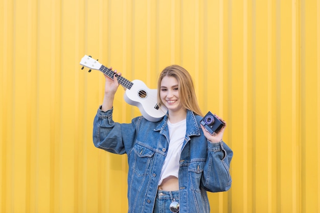 Elegante, feliz mujer inconformista está de pie con una vieja cámara y ukelele en sus manos