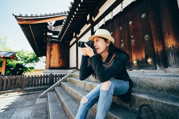 elegante feminino lente homem turista hobby amor fotografar pela câmera slr. jovem sentada nas escadas de pedra em frente ao edifício de madeira em estilo japonês antigo em dia ensolarado sob a sombra. viagem em jp.