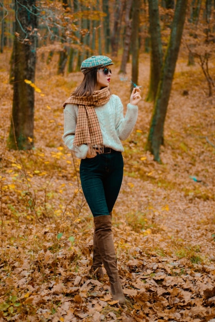 Elegante femme fatale vistiendo ropa de moda y fuma un cigarrillo en el fondo del bosque de otoño se