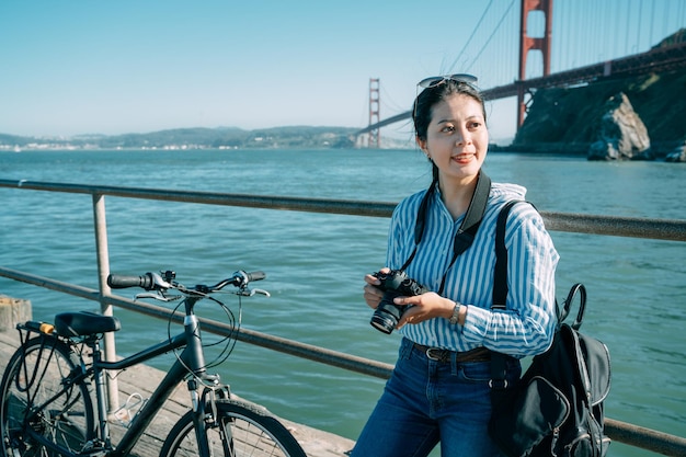 Una elegante fotógrafa asiática que viaja con una bicicleta mira a lo lejos mientras se apoya en la barandilla para tomar un descanso en un puerto deportivo en San Francisco, Estados Unidos.