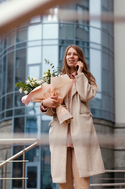 Foto elegante frau, die blumenstrauß draußen hält und smartphone verwendet