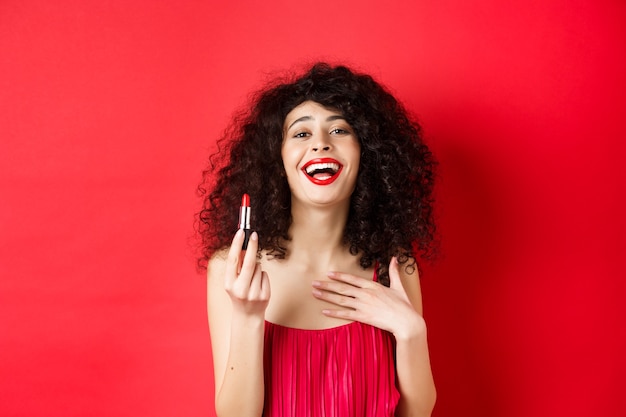 Elegante Frau im Kleid, zeigt roten Lippenstift und lachend, über Studiohintergrund stehend. Speicherplatz kopieren