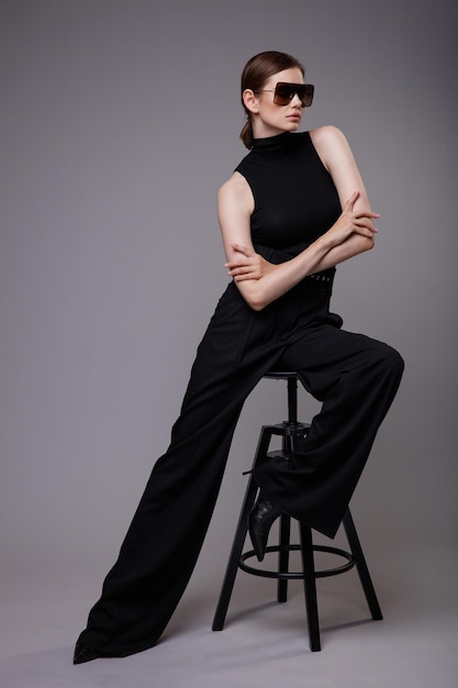 Elegante Frau in hübschen schwarzen Hosen ärmellose Top-Sonnenbrille Grauer Hintergrund Model sitzt auf Stuhl