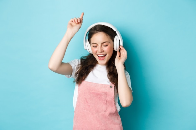 Elegante garota milenar dançando com roupa de primavera, ouvindo música em fones de ouvido e curtindo o som, de pé sobre um fundo azul.