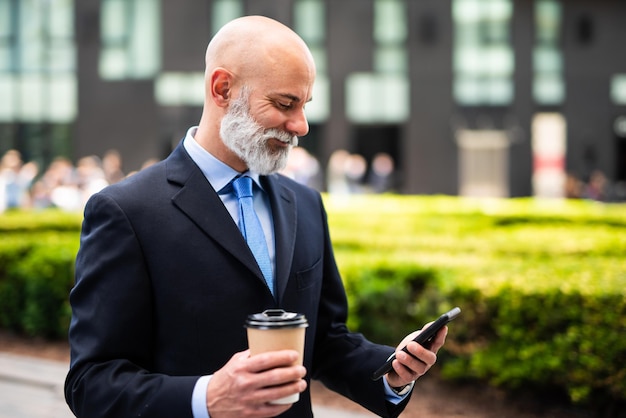 El elegante gerente senior calvo de barba blanca usa su teléfono inteligente al aire libre mientras bebe un café