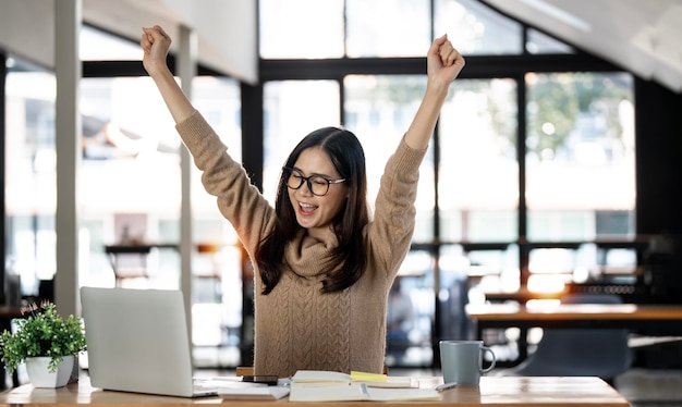 Elegante Geschäftsfrau, die im Büro mit digitalem Tablet sitzt Aufgeregte asiatische Geschäftsfrau, die die Hände hebt, um zu gratulieren, während sie am Laptop im Büro arbeitet